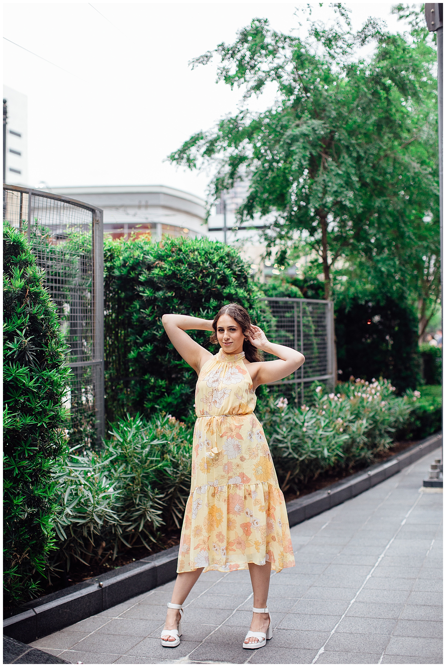 high school senior girl standing with arms behind her head in yellow floral dress by green florals in downtown Houston senior pictures