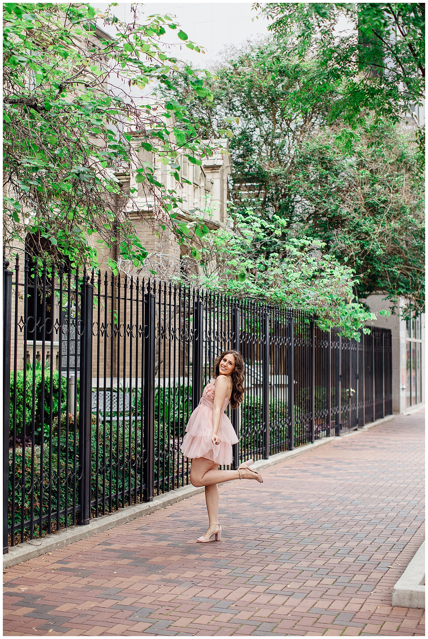 high school senior girl in pink dress standing with heel in the air in front of black iron gates in downtown Houston senior pictures