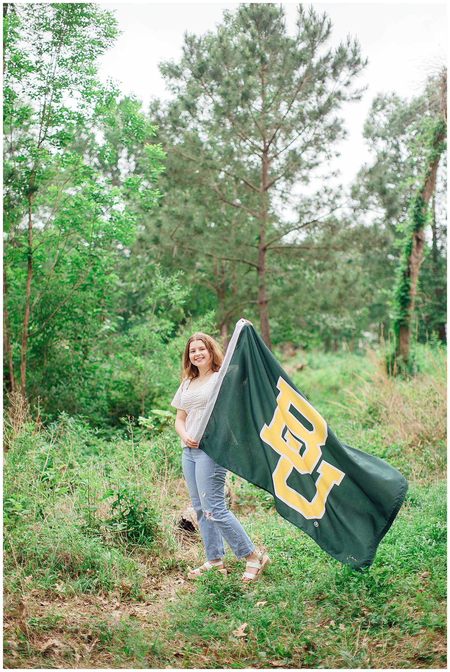 high school senior girl in jeans and white shirt standing in a green field at Houston Arboretum holding a large Baylor banner for outdoor spring senior photos