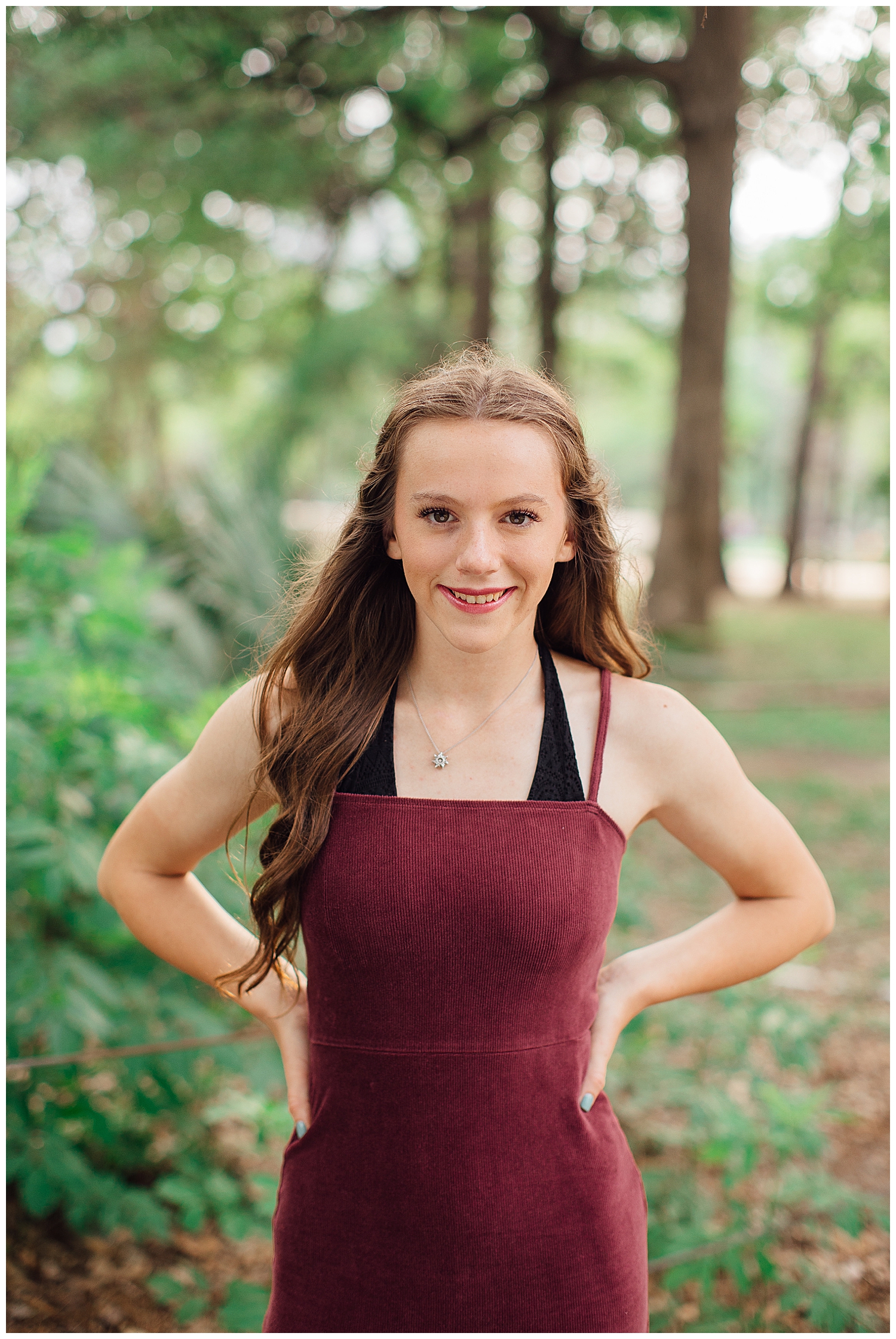 senior girl in maroon dress standing and smiling with hands on hips outdoors in Hermann park