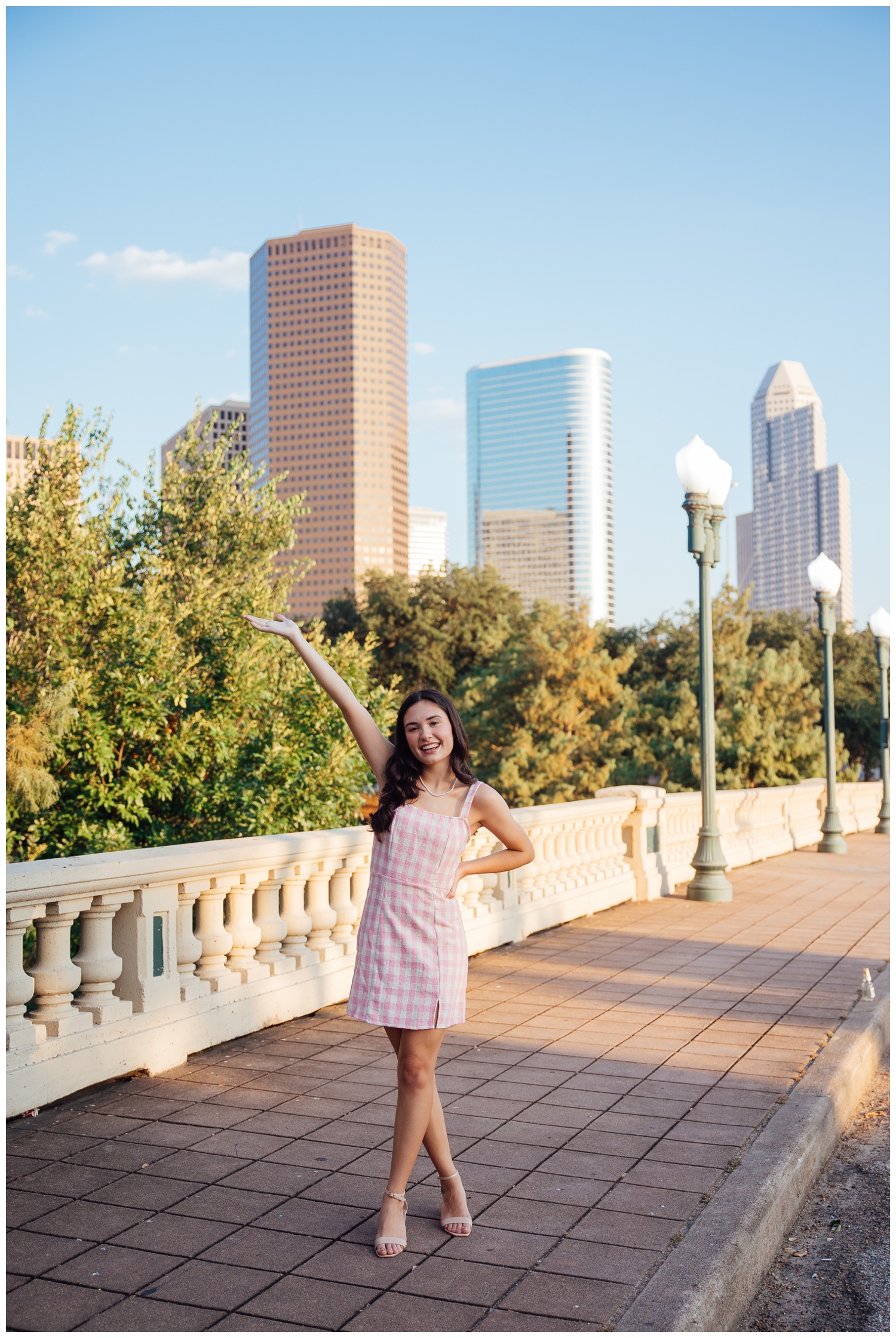 senior photos Houston skyline with girl in checkered pink dress standing with hand in air
