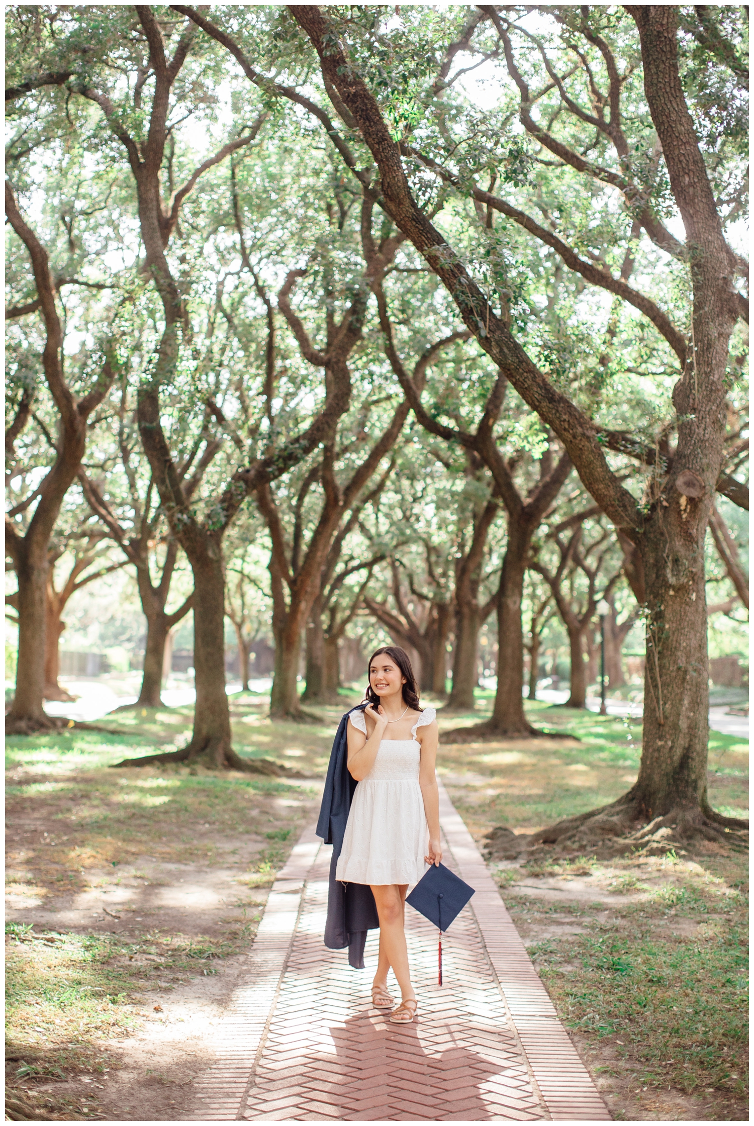 girl standing on sidewalk between trees in white dress holding cap and gown for senior photos Houston texas