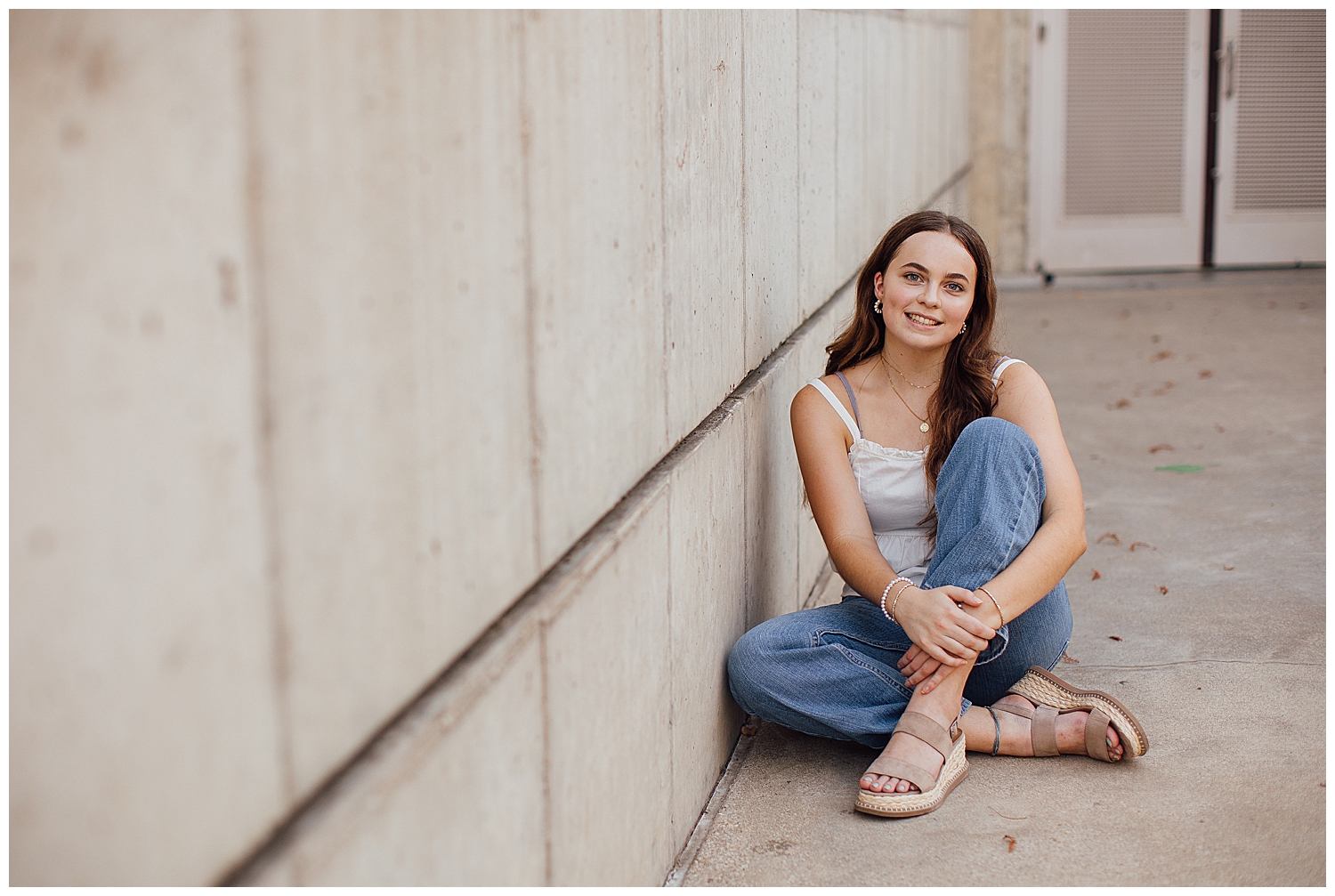 girl in jeans and white shirt sitting against cream wall Houston outdoor senior photos