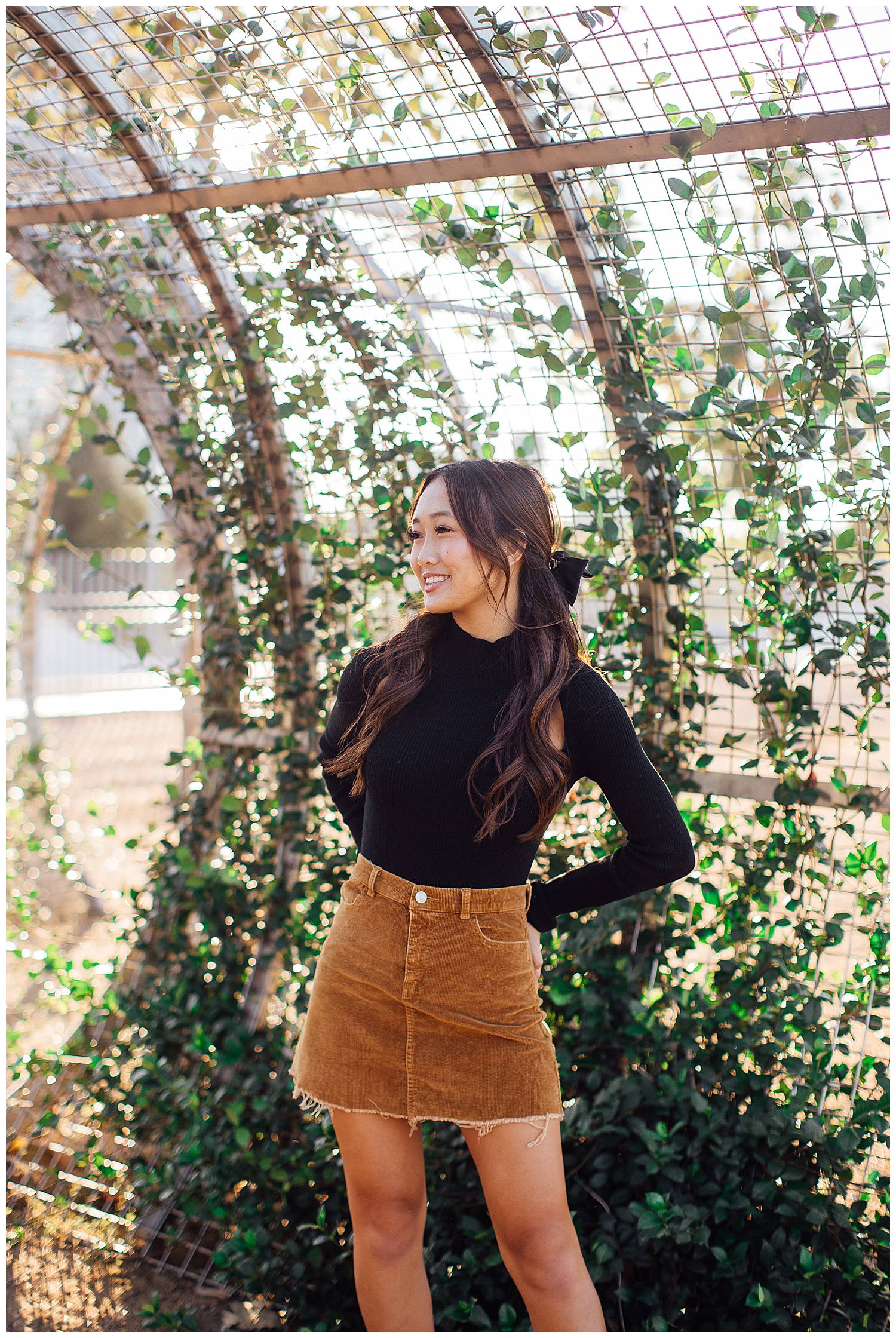 high school senior girl in black sweater and brown skirt standing in front of ivy garden arch downtown Houston