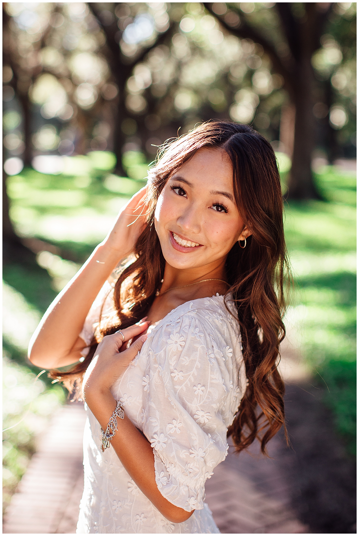 high school senior girl smiling with hand in hair in white sundress standing between trees