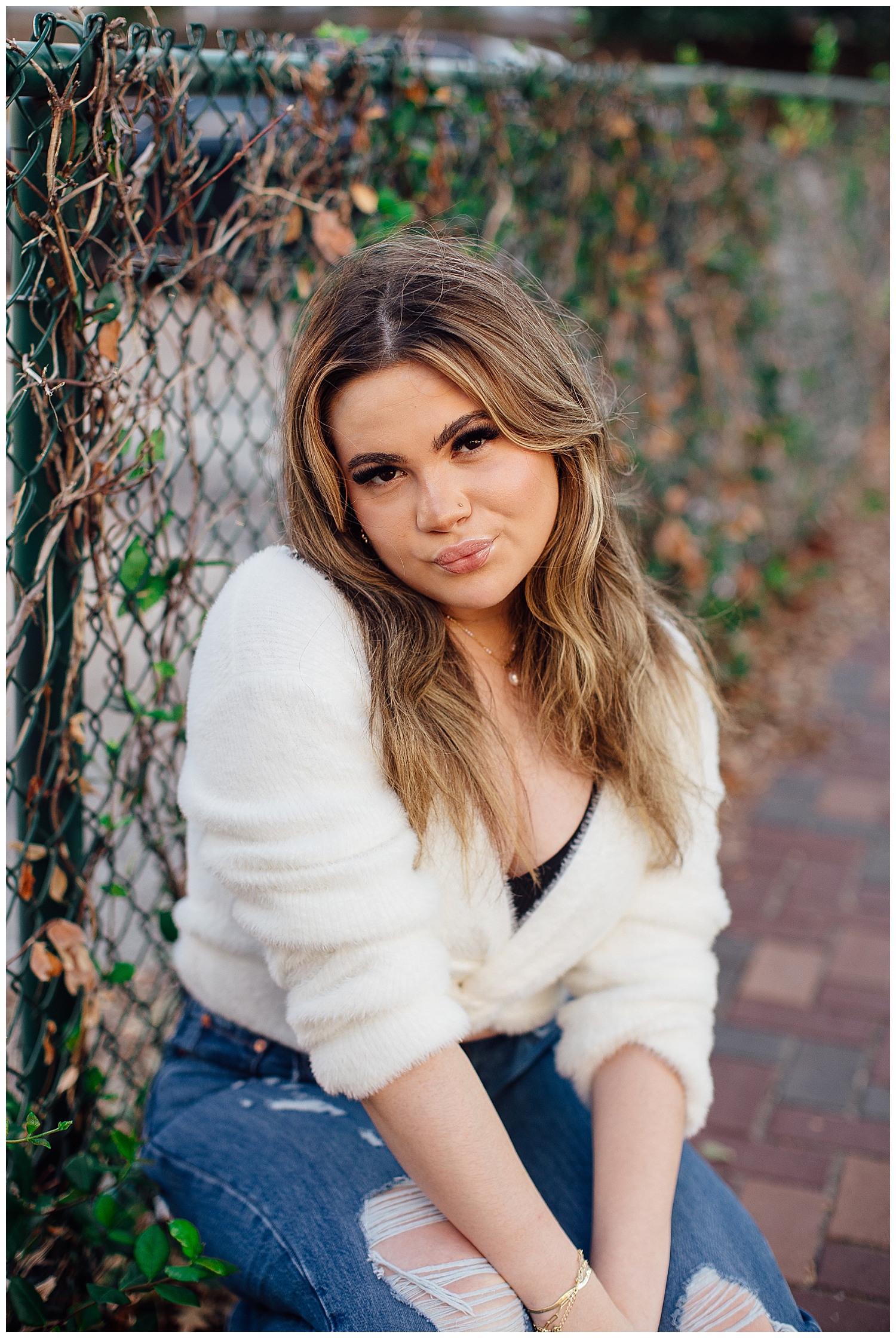 high school senior girl sitting in front of a fence in cream shirt and denim jeans