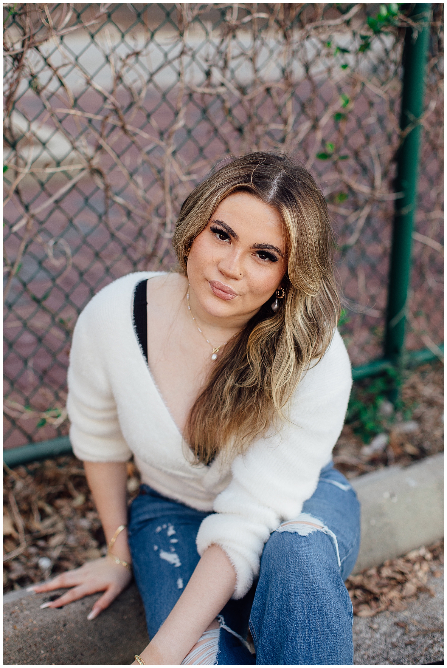 high school senior girl in cream top and jeans sitting against a fence Houston downtown senior photos