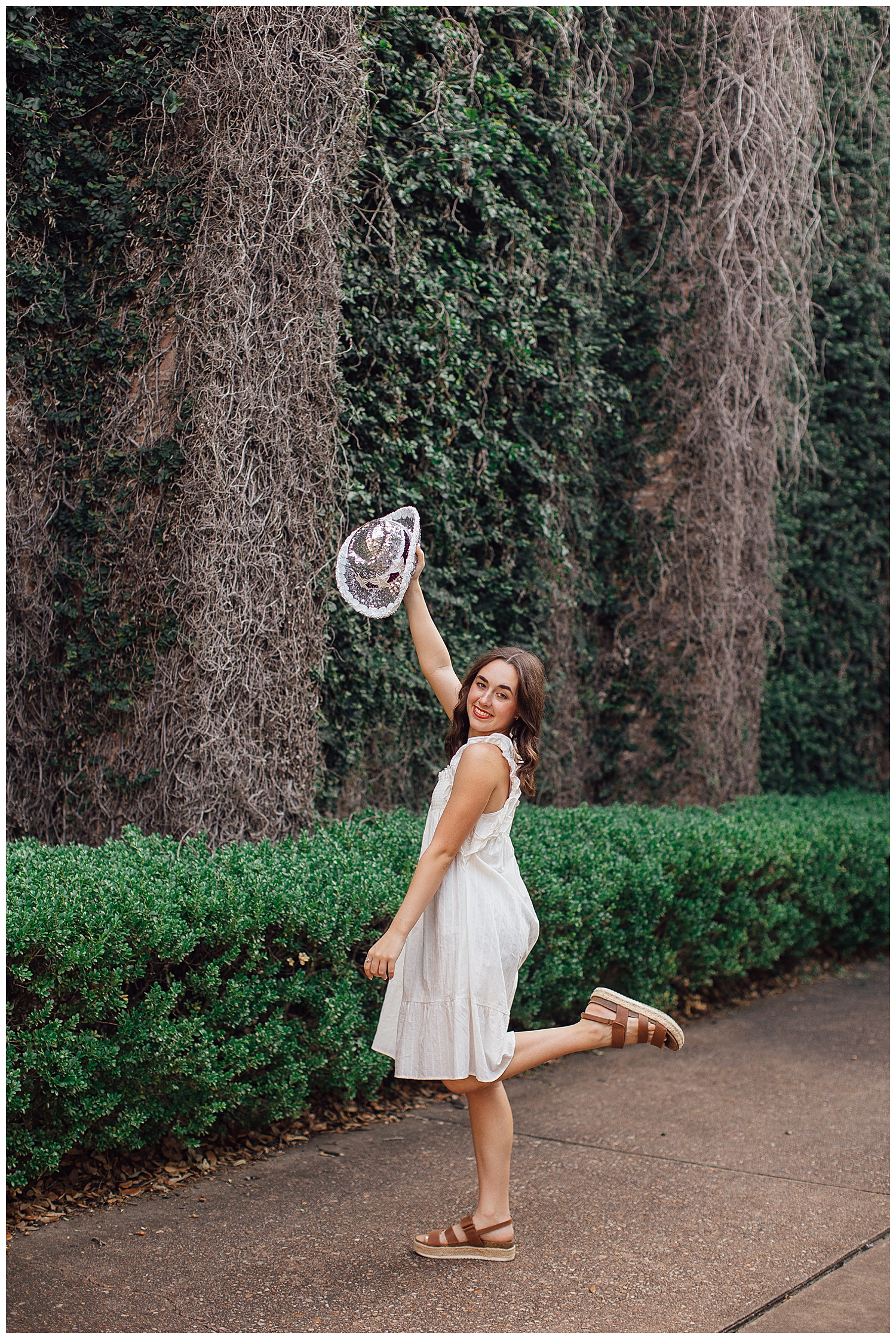high school senior girl in white dress holding drill team hat in air with leg kick in front of ivy wall Houston. Texas