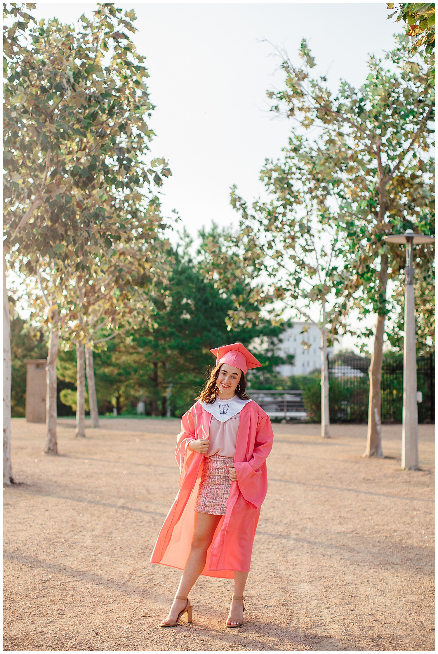 Houston skyline senior photos with girl in pink cap and gown walking by treeline