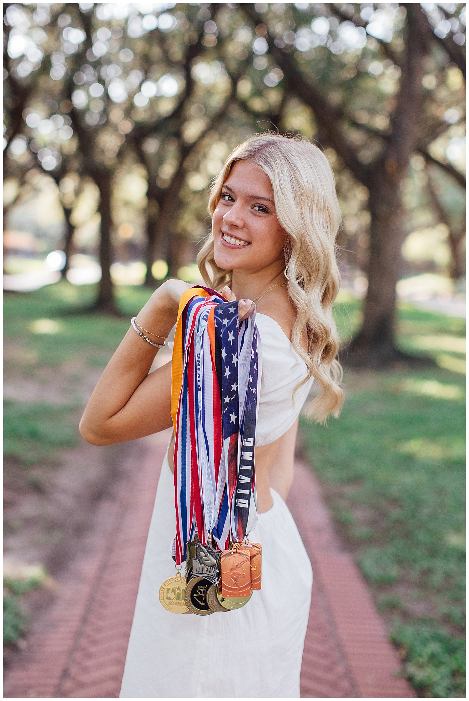 high school girl holding diving medals over her shoulder outdoors and smiling for fun senior photos Houston treeline