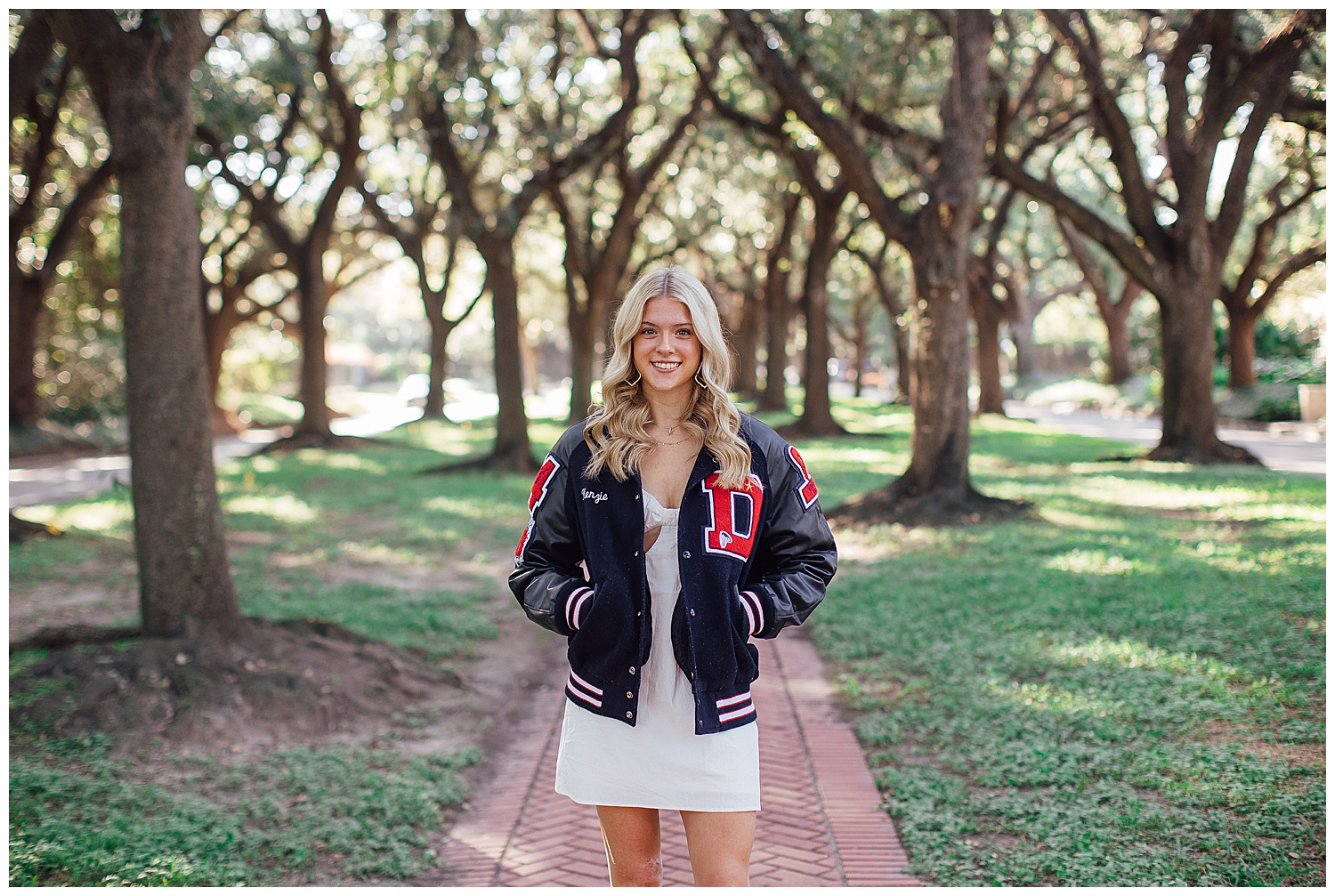 fun senior photos Houston tree line with girl wearing letterman jacket and standing on sidewalk