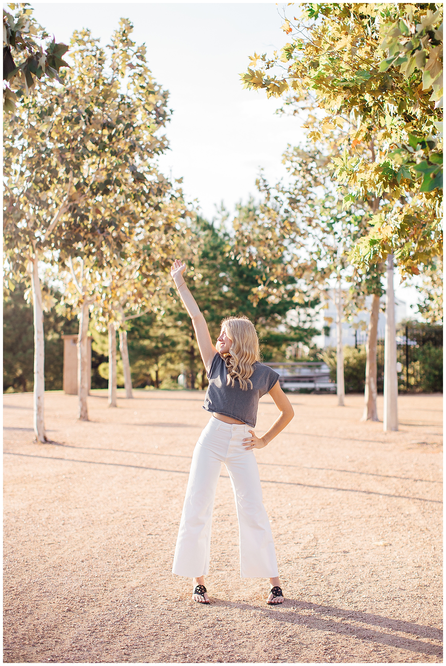 high school senior girl standing between trees Buffalo Bayou white pants gray shirt hand in air