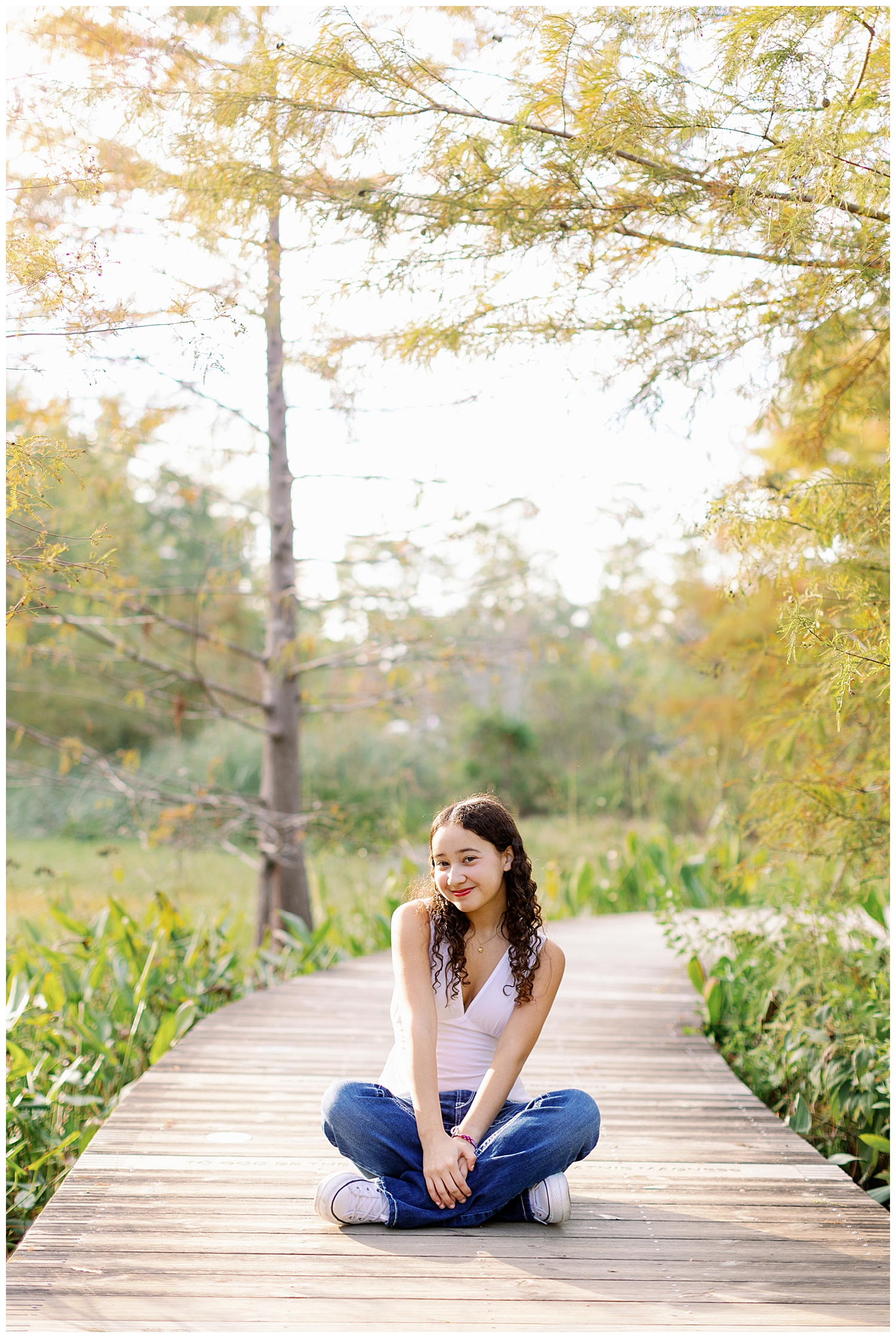 girl in white shirt and jeans sitting crisscross on boardwalk at Houston Arboretum