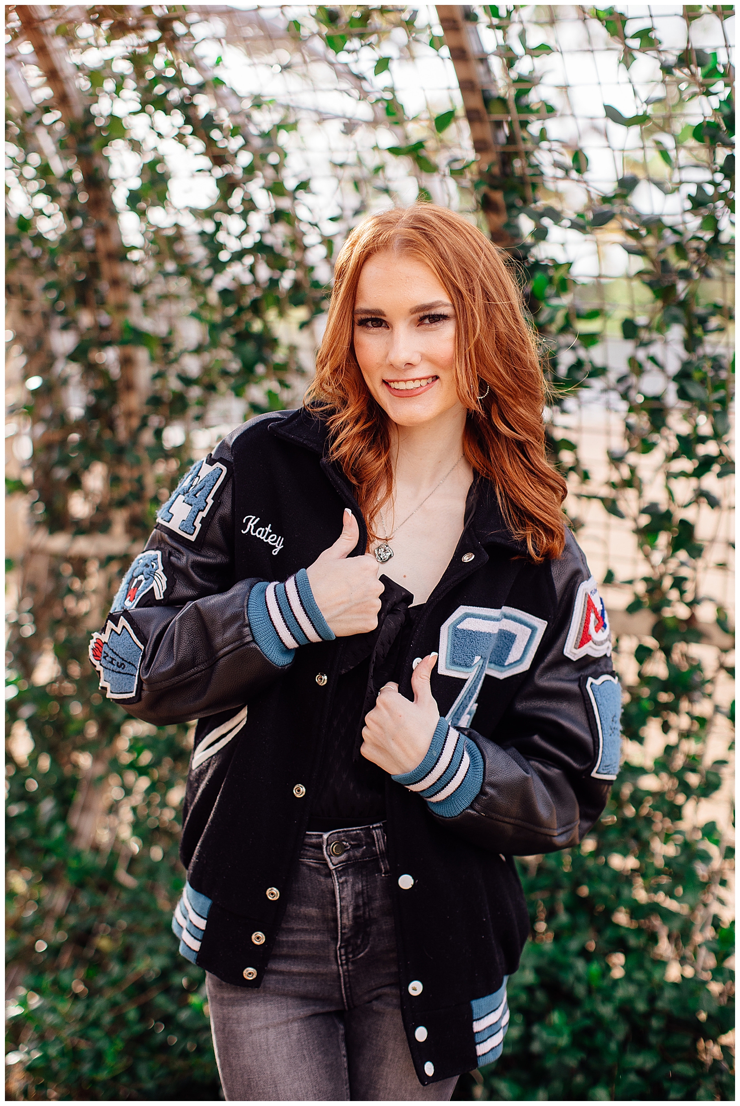 high school senior girl wearing navy letter jacket standing in front of ivy arch