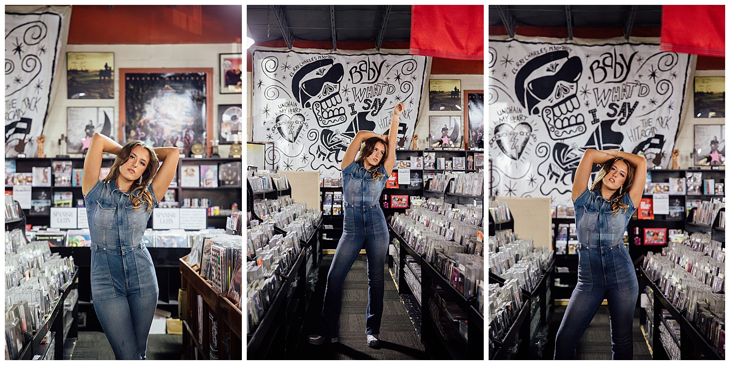 high school senior girl in denim jumpsuit standing poses with arms over head in aisle of record store