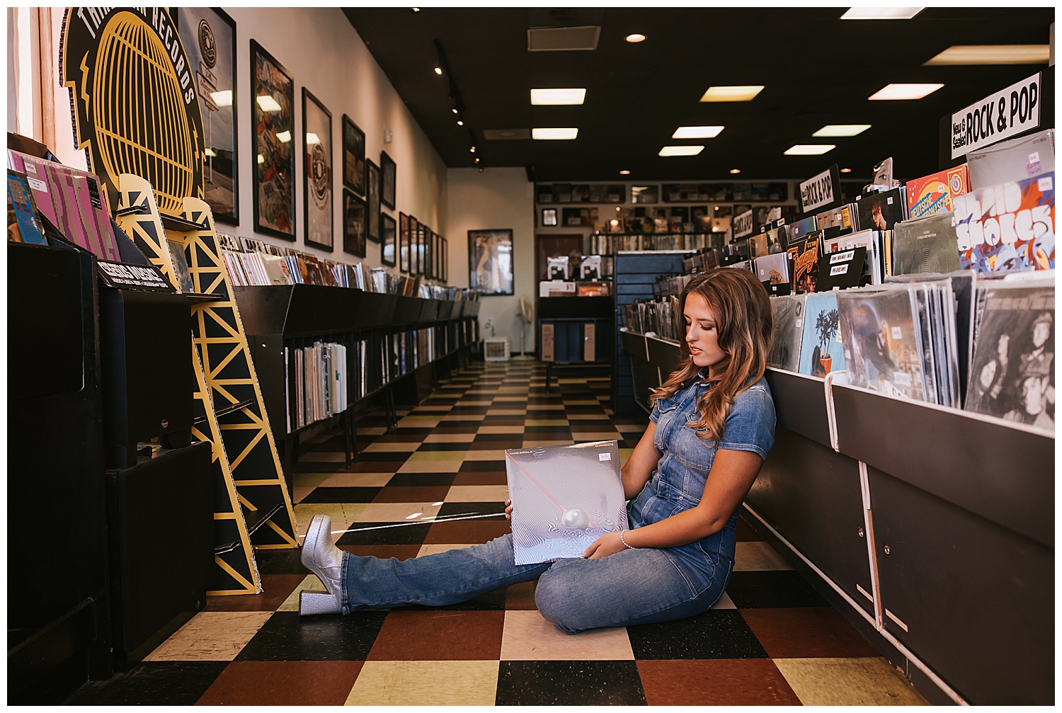girl in denim jumpsuit sitting on checkered floor holding record album inside record shop