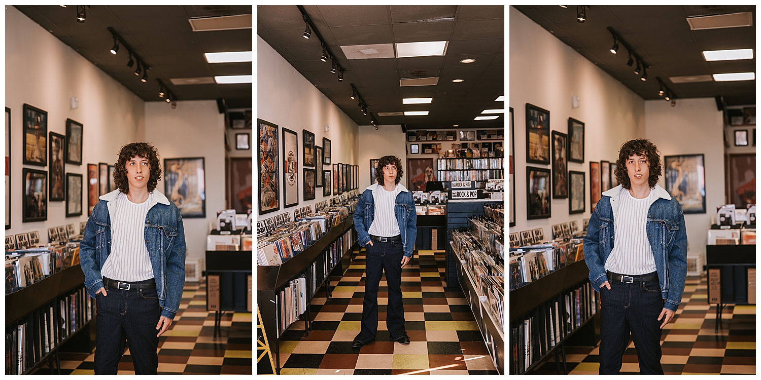 high school senior boy standing in aisle of record shop in black jeans white shirt and denim jacket
