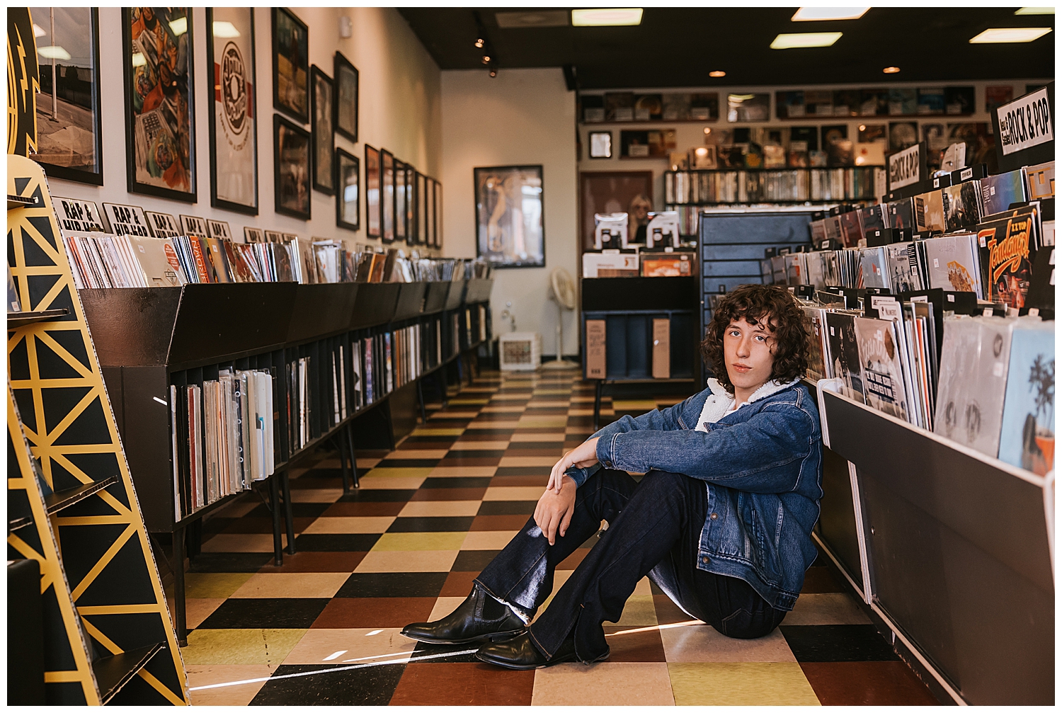 high school senior guy sitting in black jeans and denim jacket on checkered floor inside Cactus Record store Houston