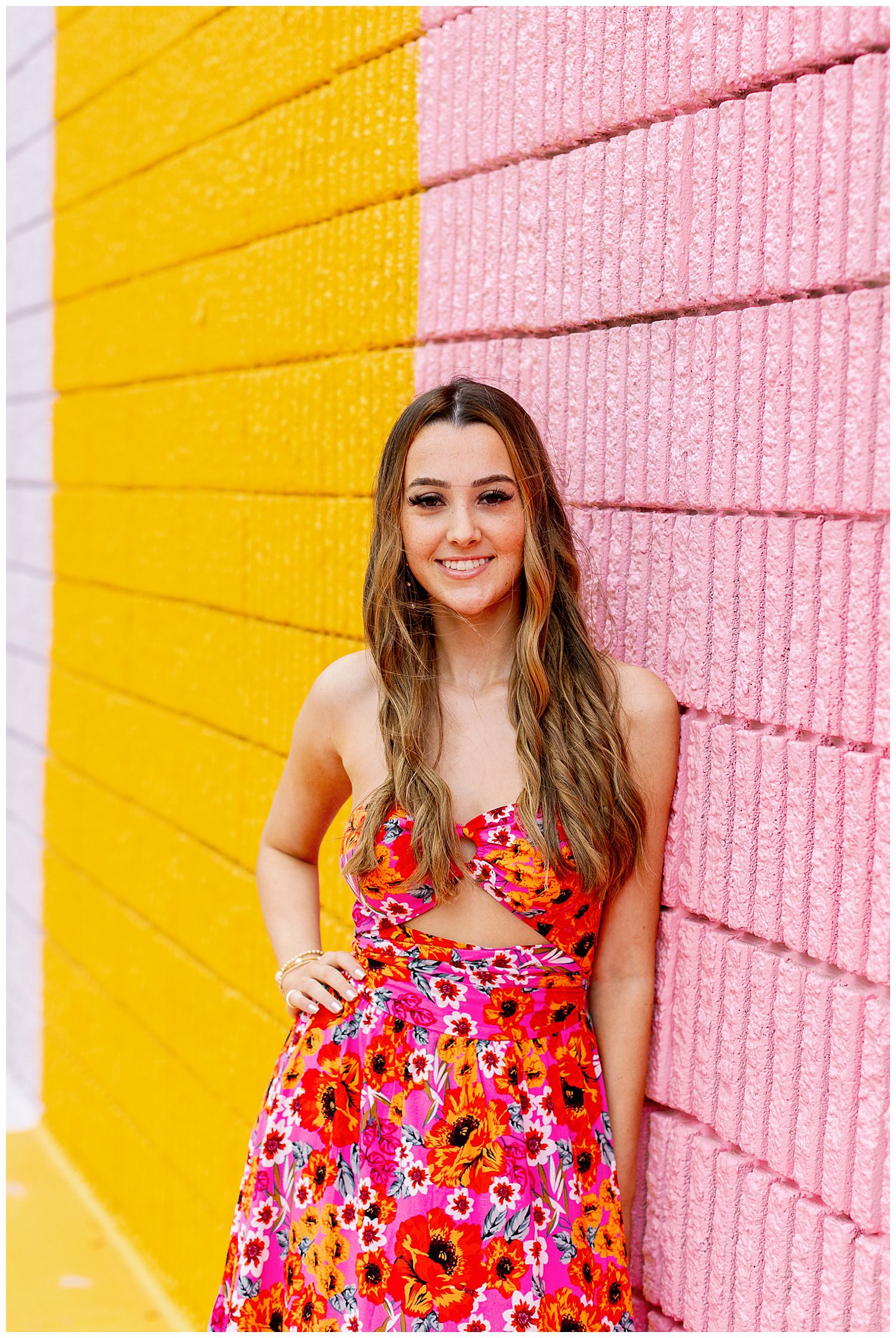 Sugar and Cloth Color Wall portraits of a girl in red floral dress smiling with hands on hip leaning on pink wall