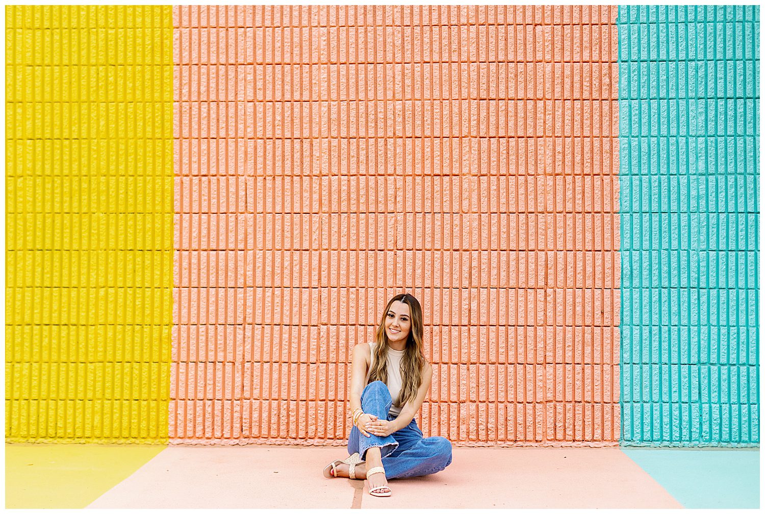 high school senior girl in jeans and beige shirt sitting with legs crossed in front of Sugar and Cloth Color Wall portraits in Houston Texas
