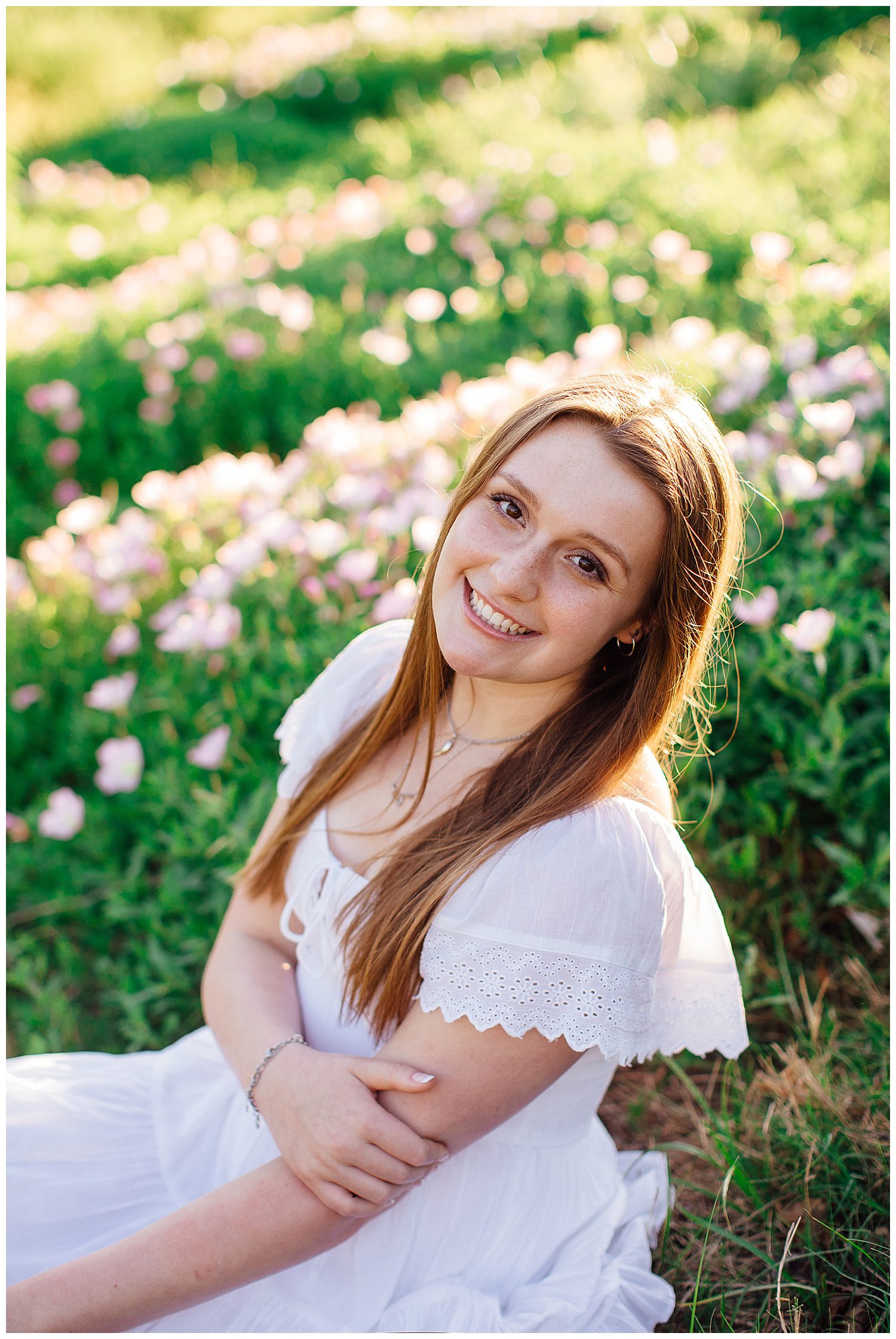 high school senior girl in white sundress leaning toward camera sitting in green field with pink flowers downtown Houston