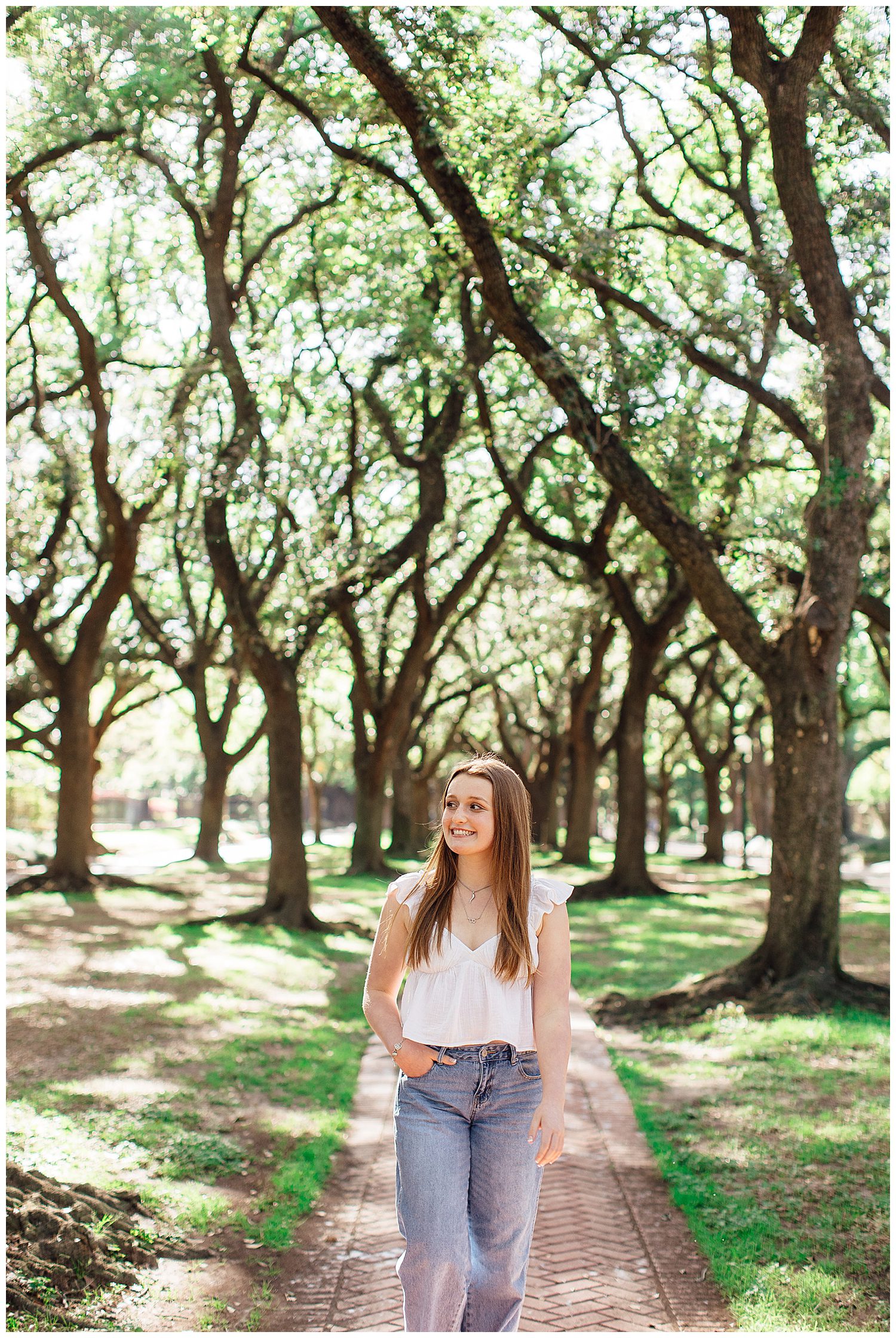 high school senior girl walking with hand in pocket wearing jeans and white blouse outdoors by trees Houston