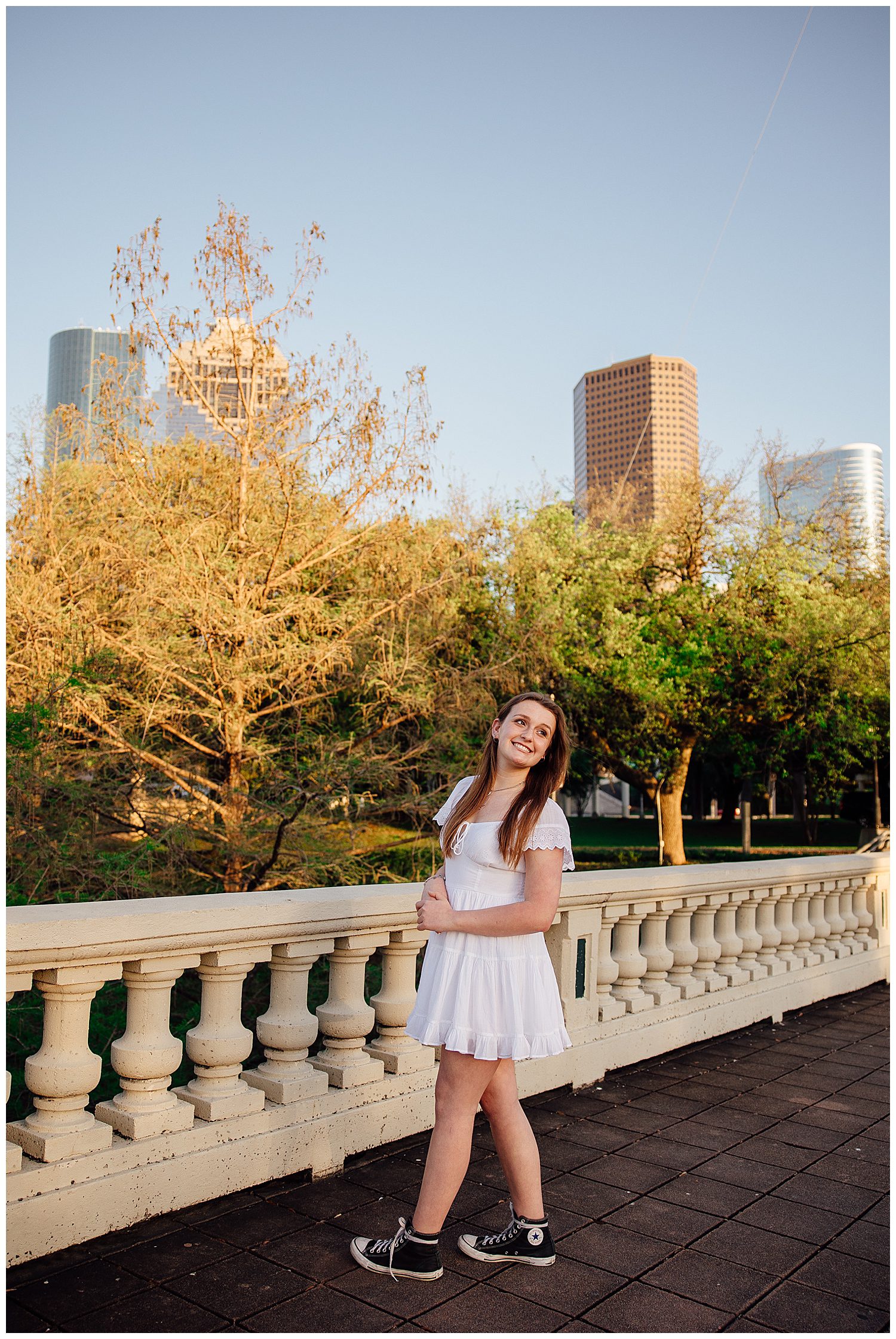 senior girl standing On Sabine Street bridge in front of Houston skyline wearing white sundress looking over her shoulder