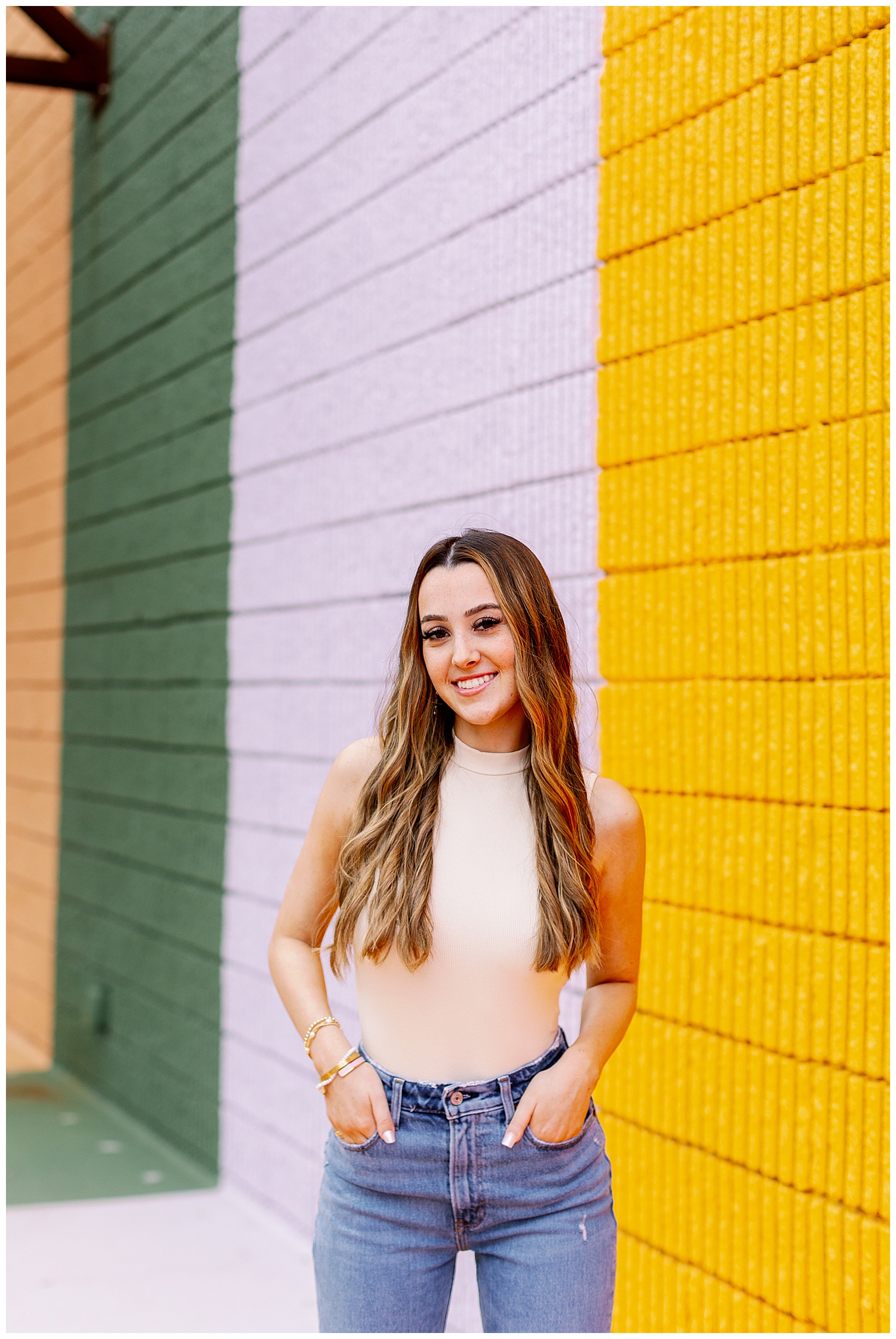girl standing with hands in jeans smiling at camera in front of multicolored wall