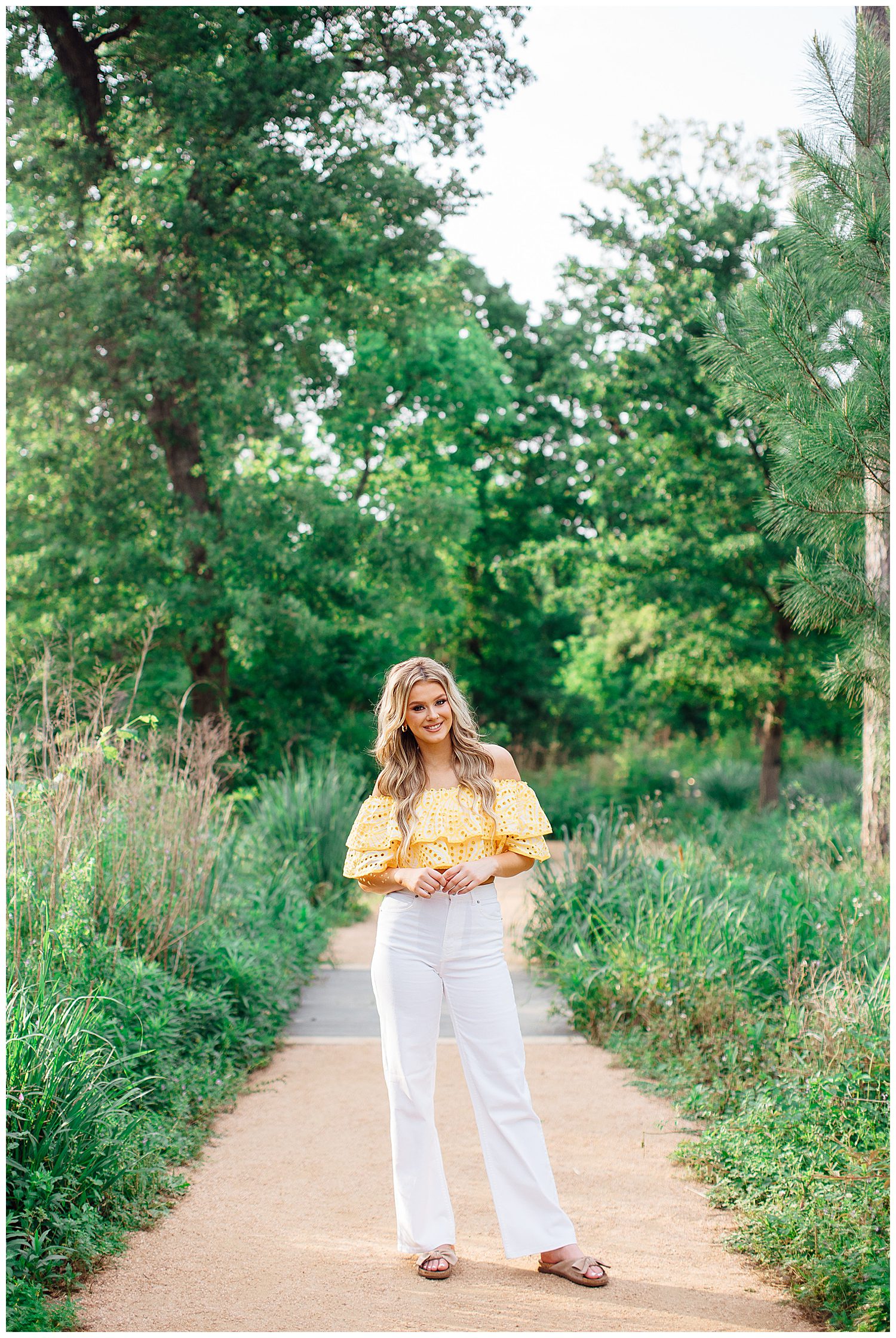 girl in white pants and yellow shirt standing outdoors on pathway Houston Arboretum colorful spring senior pictures